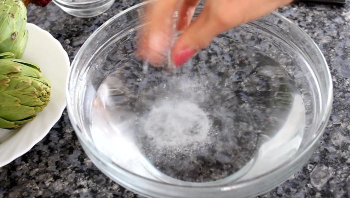 Preparar las alcachofas: Lavarlas y colocarlas en un bol con agua muy fría y un puñado de sal para evitar que se oxiden. Cortar el tronco, quitar las hojas exteriores duras y la parte superior más dura del corazón. Partir por la mitad y retirar los pelillos del interior.